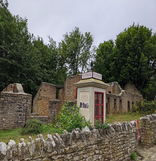 A telephone box stands in front of some derelict brick buildings without a roof, in front of green trees in the abandoned village